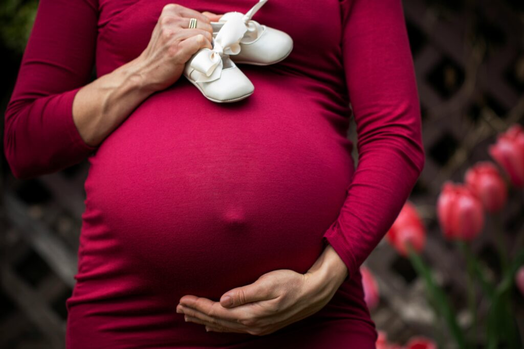 A pregnant woman in a burgundy dress is holding her belly with her left hand and a pair of white baby shoes in her right hand