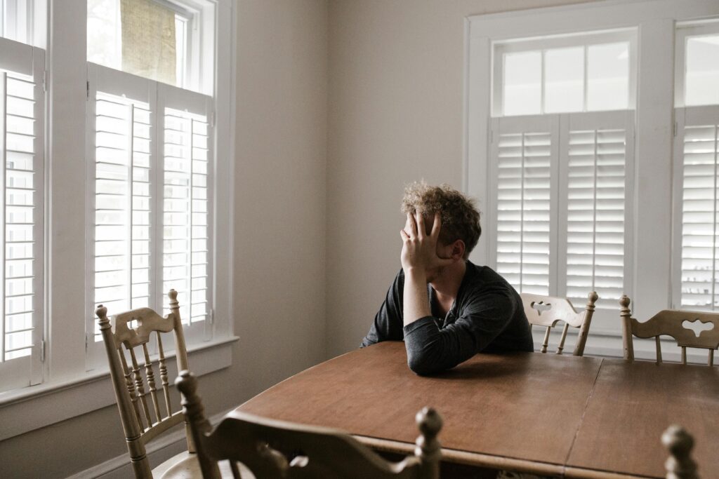 A sad man sitting on a chair in front of a window, covering his face with his arm on the wooden table