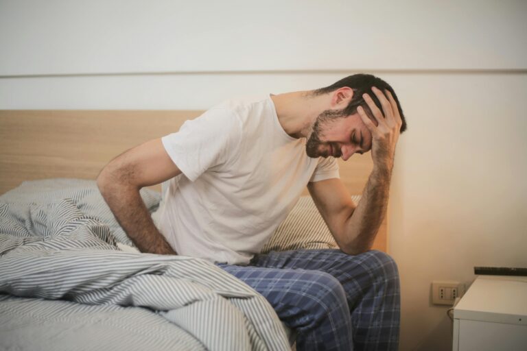 A young man sitting on a bed, writhing in pain, holding his head with one hand and his hip with the other