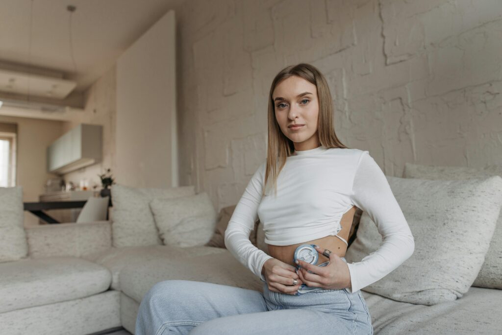 A young woman injects medication into her abdomen while sitting on a white couch in a room with white furniture